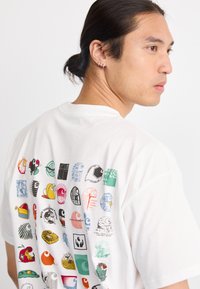 Camiseta blanca de algodón con un impresionante estampado gráfico en la parte trasera, que presenta diversos motivos de dibujos animados y patrones en múltiples colores y estilos.