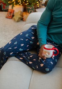 Blauwe pyjamabroek met feestelijke patronen, waaronder zuurstokken en peperkoekmannetjes, gecombineerd met een groen shirt met lange mouwen en een kerstmanmok.