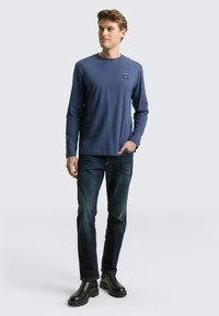 Granatowa koszulka z długim rękawem i okrągłym dekoltem, z małą naszywką z logo na piersi, zestawiona z ciemnymi dżinsami i czarnymi butami.