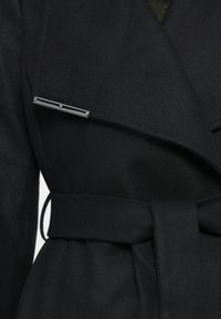Casaco preto envolvente com uma textura suave, apresentando um detalhe em barra prateada e um cinto a condizer. Vista em close que destaca os pormenores do design.