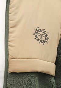 Μπεζ εξωτερικό μπουφάν με επένδυση από απαλό πράσινο φλις. Διαθέτει κέντημα ήλιου στο μπροστινό μέρος και αντίθετο εσωτερικό τελείωμα σε ελιά απόχρωση.