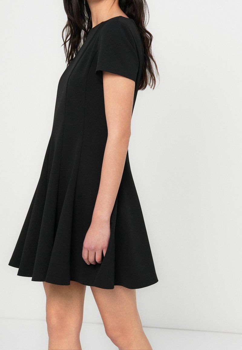 Femme portant une robe noire à manches courtes en ligne A avec une jupe évasée, debout de profil sur un fond clair uni.