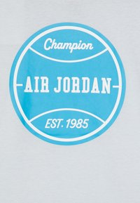 Szara koszulka z niebieskim okrągłym logo, na którym widnieją napisy "Champion", "AIR JORDAN" oraz "EST. 1985" w białym kolorze. Gładki materiał bawełniany.