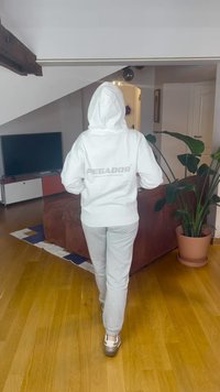 Sweatshirt branca com capuz com o logótipo "PEGADOR" nas costas, combinado com calças de fato de treino cinza claro e sapatos metálicos prateados. Chão de madeira e plantas de interior.