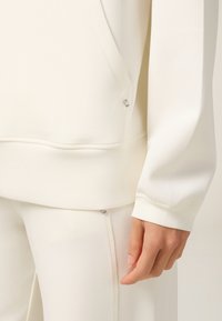 Sudadera blanca de tela suave con detalle de bolsillo lateral y textura lisa. Presenta herrajes plateados cerca de los cremalleras. Diseño ajustado.