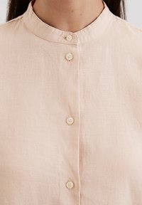 Lichtroze linnen blouse met een staande kraag en zes knoopsluitingen. De stof heeft een licht gestructureerde afwerking. De knopen zijn crème kleurig.