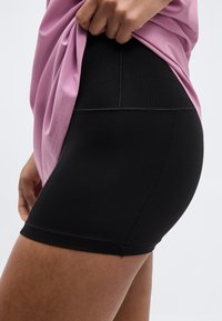 Άτομο σηκώνει ροζ μπλουζάκι για να δείξει μαύρα, ψηλόμεσα, εφαρμοστά σορτς πάνω από τον μηρό και στο ισχίο, σε απλό φόντο.