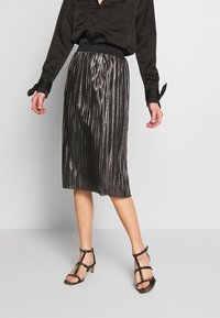 Jupe plissée métallique noire avec une ceinture, associée à un chemisier en satin noir et des talons noirs à brides. Design longue longueur au sol.