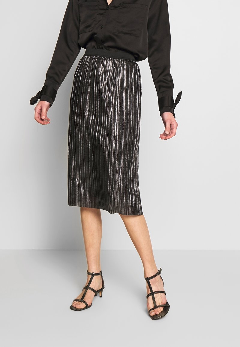 Jupe plissée métallique noire avec une ceinture, associée à un chemisier en satin noir et des talons noirs à brides. Design longue longueur au sol.