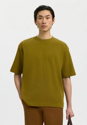 Giovane uomo con capelli neri lisci che indossa una maglietta oversize verde oliva e pantaloni marroni, tiene una borsa con tracolla marrone scuro nella mano destra.