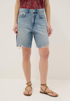 Femme portant un short en jean bleu clair déchiré, un haut rouge à motifs, et des sandales plates marron à brides, debout devant un fond uni.