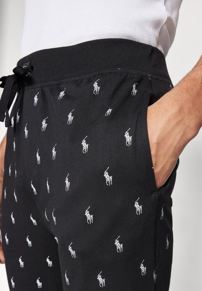 Pantalons de survêtement noirs avec une taille ajustée, arborant un motif répétitif de logo de joueur de polo blanc et des poches latérales. Texture de tissu douce.