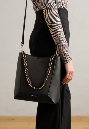 Melnas ādas plecu soma ar zelta ķēdes akcentu, stūrainu dizainu, gludu tekstūru un uzrakstu "ANNA FIELD" priekšpusē.