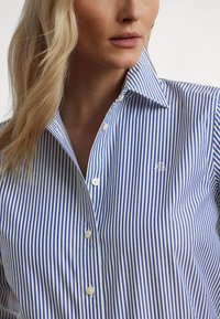 Blå og hvitstripet knappeskjorte med spiss krage og brodert logo på brystet; bomullsstoff med glatt tekstur.