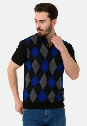Mann mit kurzen dunklen Haaren und Bart, der einen ärmellosen Pullover mit blauem, grauem und schwarzem Rautenmuster über einem schwarzen kurzärmeligen Hemd und blauen Jeans trägt.
