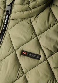 Olijfgroen gewatteerde jas met een zwarte rits en een rechthoekig zwart label met "SUPERDRY" in rood en wit. Geweven materiaal.