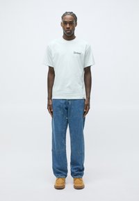 Hombre de pie frente a un fondo blanco, vestido con una camiseta blanca de Dickies, jeans azules holgados con una costura frontal única y botas de trabajo color beige.