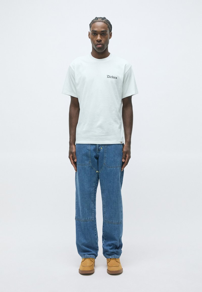Man staat tegen een witte achtergrond, gekleed in een witte Dickies T-shirt, losse blauwe jeans met een unieke voornaad, en tan kleurige werkschoenen.