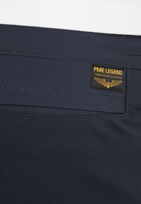 Marineblauwe broek met een soepele textuur, een achterzak en een merklabel met gouden tekst en een gevleugeld logo.