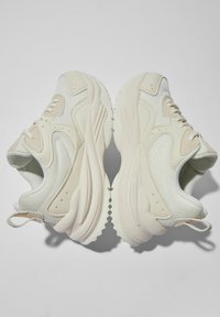 Beżowe buty sportowe z teksturowaną siateczką, zaokrąglonym noskiem, grubą gumową podeszwą i pętelką na pięcie. Krzywe akcenty wzornicze wzdłuż boków.