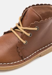 Hnědá kožená kotníková bota s vlněným okrajem, světle hnědé šňůrky provlečené kovovými očky a prošitá gumová podrážka.