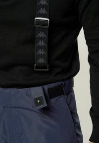 Crne naramenice s ponovljenim uzorkom loga preko crnog džempera, pričvršćene za tamnoplave hlače s dugmetom na pritiskanje i čičak zatvaranjem.