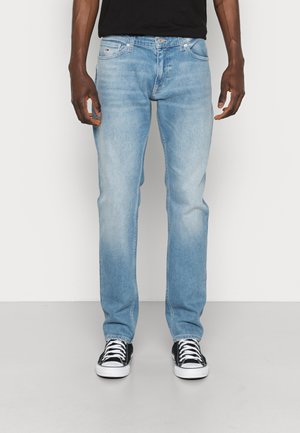 Mann trägt hellblaue Jeans mit geradem Bein, schwarzes T-Shirt und schwarze Low-Top-Sneakers mit weißen Schnürsenkeln und Sohlen, steht auf grauem Boden.