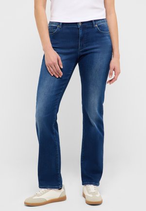 Kobieta w ciemnoniebieskich dżinsach typu bootcut, białej bluzce i białych trampkach stoi na tle jednolitego jasnego tła.