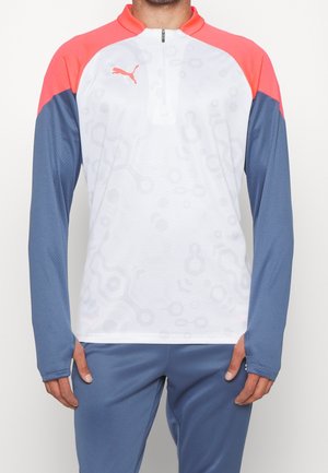 Man draagt een sporttop met lange mouwen met een wit lijf, koraalkleurige schouders en blauwe mouwen, gecombineerd met een blauwe broek, staand tegen een effen achtergrond.