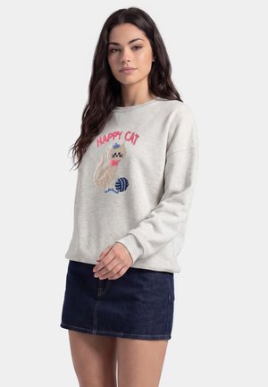 Ung kvinde iført en lysegrå sweatshirt med en kat og teksten "Happy Cat", kombineret med et mørkeblåt cowboyskørt, stående på en ensfarvet baggrund.