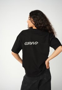 Mujer con cabello rizado oscuro que lleva una camiseta negra con "anno" bordado en la parte de atrás, combinada con pantalones negros, de pie con las manos en las caderas.