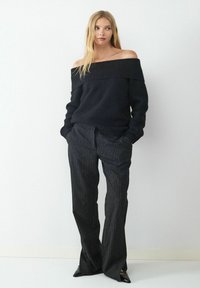 Schwarzer schulterfreier Pullover mit geripptem Kragen, kombiniert mit gestreiften Hosen. Die Hosen haben ein gerades Bein und einen subtilen Glanz.