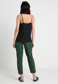 Top noir à bretelles avec un pantalon vert ajusté, présentant une coupe ample et des ourlets retroussés. Des chaussures noires plates complètent la tenue.