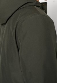 Veste à capuche vert foncé en tissu lisse et léger. Présente une coupe ajustée, des coutures visibles et une fermeture à pression au niveau du col.