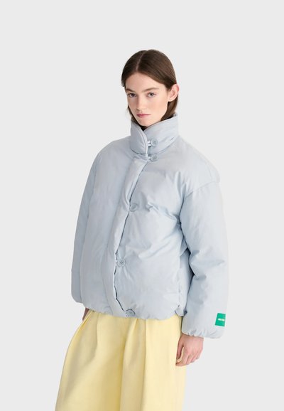 Femme aux cheveux châtain clair portant une doudoune bleue pâle boutonnée et un pantalon jaune ample sur un fond clair uni.