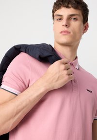 Švelnios rožinės spalvos polo marškiniai su apykakle ir dviem mygtukais, ant kurių yra tamsus logotipas. Tamsiai mėlynas švarkas numestas ant vienos peties.