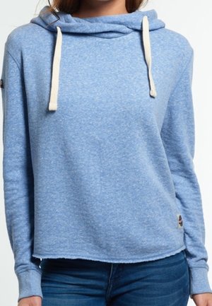 Kobieta ubrana w jasnoniebieską bluzę z kapturem i białymi sznurkami, w ciemnoniebieskich dżinsach, pokazana od ramion do bioder.