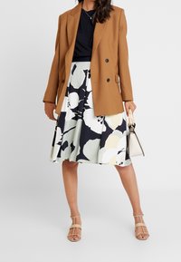 Ruskea kaksirivinapillinen bleiseri mustan topin päällä, yhdistettynä kukalliseen, polvipituiseen hameeseen, jossa on kermaan, laivastonsiniseen ja vihreään vivahtavia sävyjä, ja beigekkaisiin nauhalla oleviin sandaaleihin.