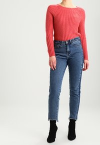 Pull rouge en maille côtelée à manches longues, associé à un jean en denim bleu taille haute avec un ourlet brut, et des bottines noires.