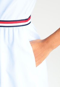 Helles blaues Kleid aus strukturiertem Stoff, mit einer taillierten Passform und einem kontrastierenden Gürtel in Marineblau, Rot und Weiß mit Streifen. Sichtbare Seiten Tasche.