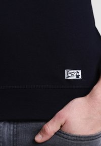 Nahaufnahme eines dunkelblauen Kleidungsstücks mit einem kleinen rechteckigen weißen Etikett, auf dem "Brooklyn NYC" steht, und einer Hand, die teilweise in der grauen Jeans-Tasche steckt.