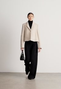Μπεζ μάλλινο σακάκι με εφαρμοστή σχεδίαση και κλείσιμο με ένα κουμπί, συνδυασμένο με μαύρο παντελόνι φαρδιάς γραμμής και μικρό μαύρο τσαντάκι χειρός.