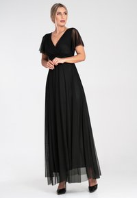 Schwarzes Maxikleid mit transparenten kurzen Ärmeln, V-Ausschnitt und fließendem Rock. Texturierte Überlagerung am Saum, kombiniert mit schwarzen spitzen Absätzen.