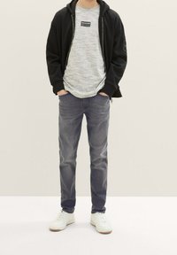 Felpa con cerniera nera sopra una t-shirt grigia a trama, abbinata a jeans grigi e sneakers bianche. Abbigliamento casual senza loghi visibili sui jeans.