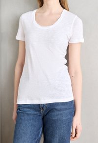 Hvid kortærmet t-shirt med scoop halsudskæring, lavet af letvægtsstof. Parret med blå denim jeans mod en ensfarvet baggrund.