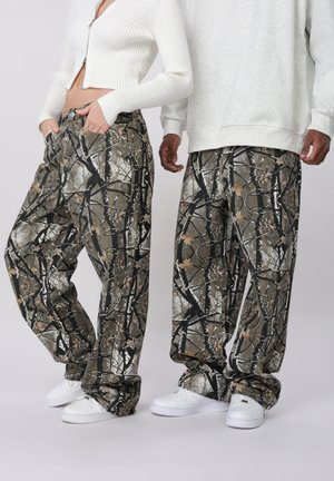 Zwei Personen tragen passende lockere Camouflage-Hosen und weiße Turnschuhe, eine mit einem weißen, gekürzten Zip-Pullover, die andere mit einem grauen Sweatshirt.