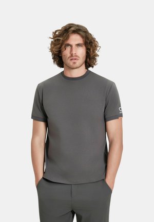 Uomo con capelli ricci che indossa una maglietta a maniche corte grigio scuro tinta unita e pantaloni abbinati, in piedi con le mani in tasca su sfondo bianco.