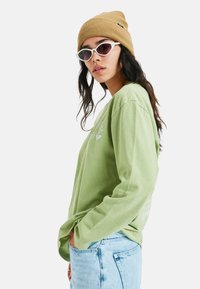 Jasnozielona koszula z długim rękawem o luźnym fasonie, w połączeniu z wyblakłymi niebieskimi dżinsami. Uzupełniają ją brązowa czapka typu beanie z prążkowanym wzorem i białe okulary przeciwsłoneczne.