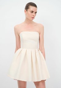 Robe crème sans bretelles avec un corsage ajusté et une jupe évasée plissée. Sa texture lisse et ses poches latérales sont des caractéristiques notables.