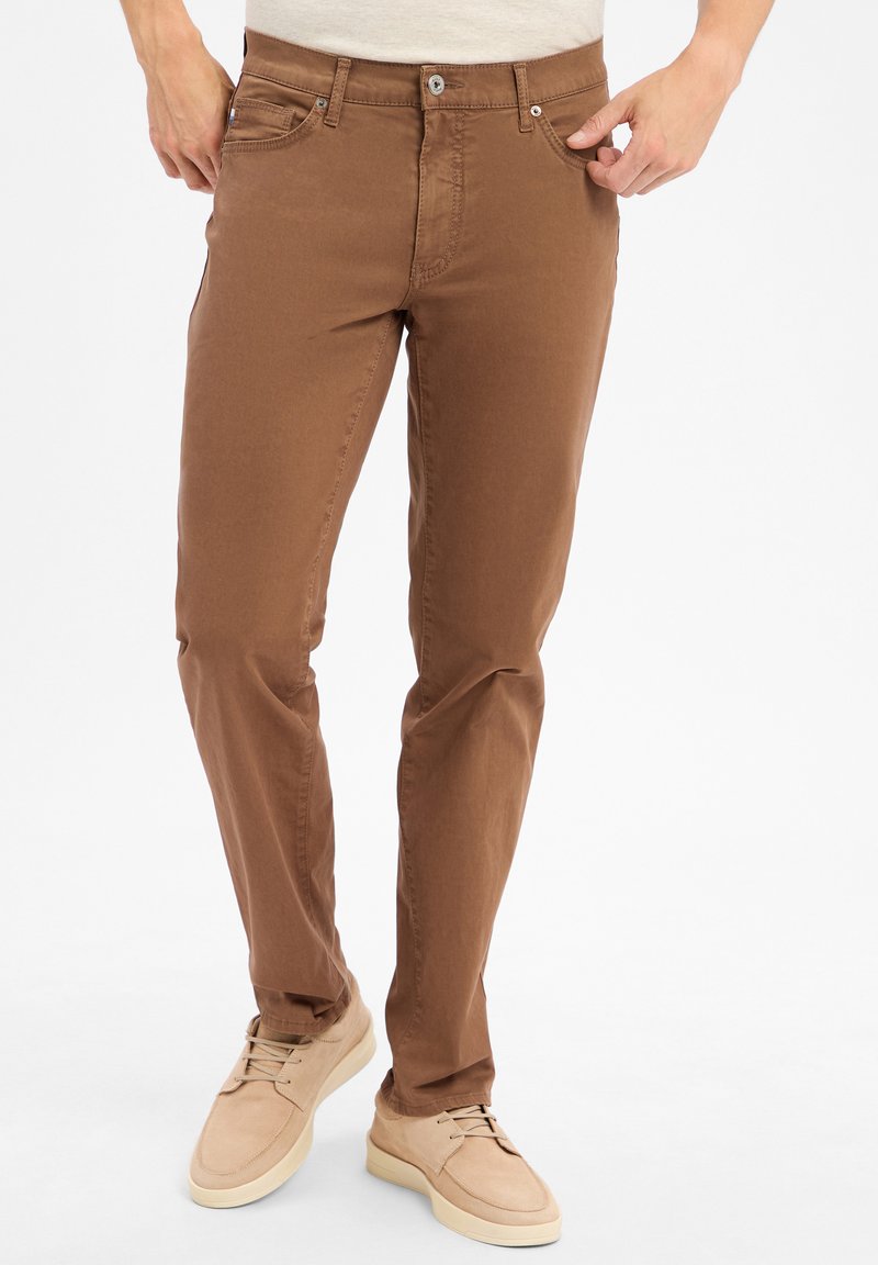 Pantalons en mélange de coton marron avec une coupe slim, dotés de poches avant, d'une fermeture éclair et d'une texture lisse. Associés à des chaussures décontractées de couleur claire.